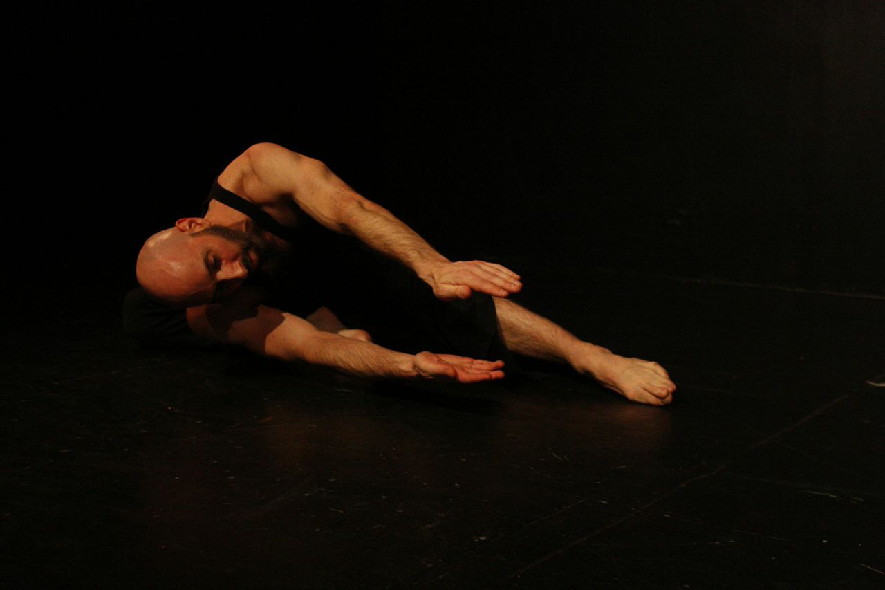 Dancer Daniel Gwirtzman lays on his right side, both of the hands extend towards the audience as if he is holding something between them